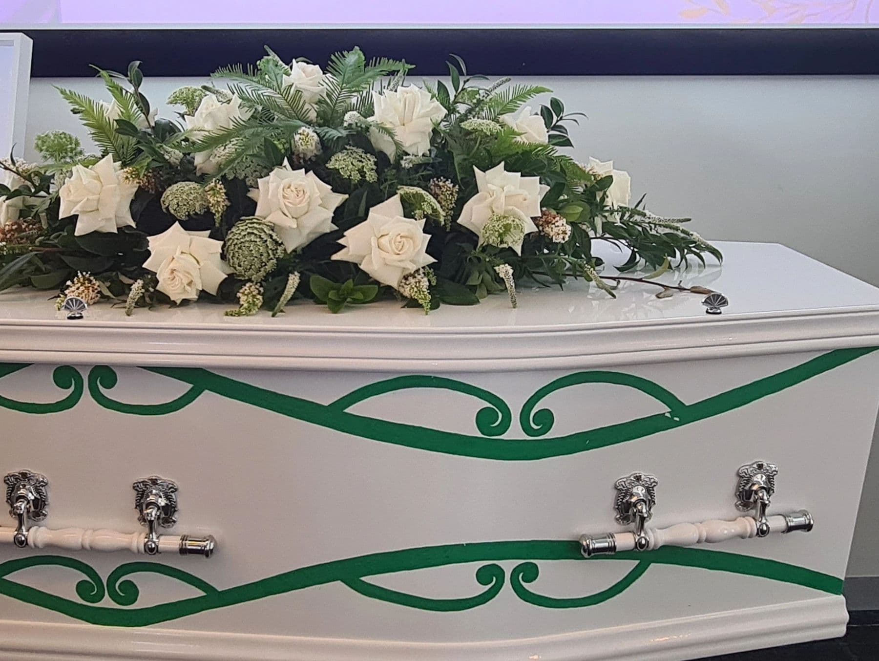 White coffin adorned with white roses and green leaves.