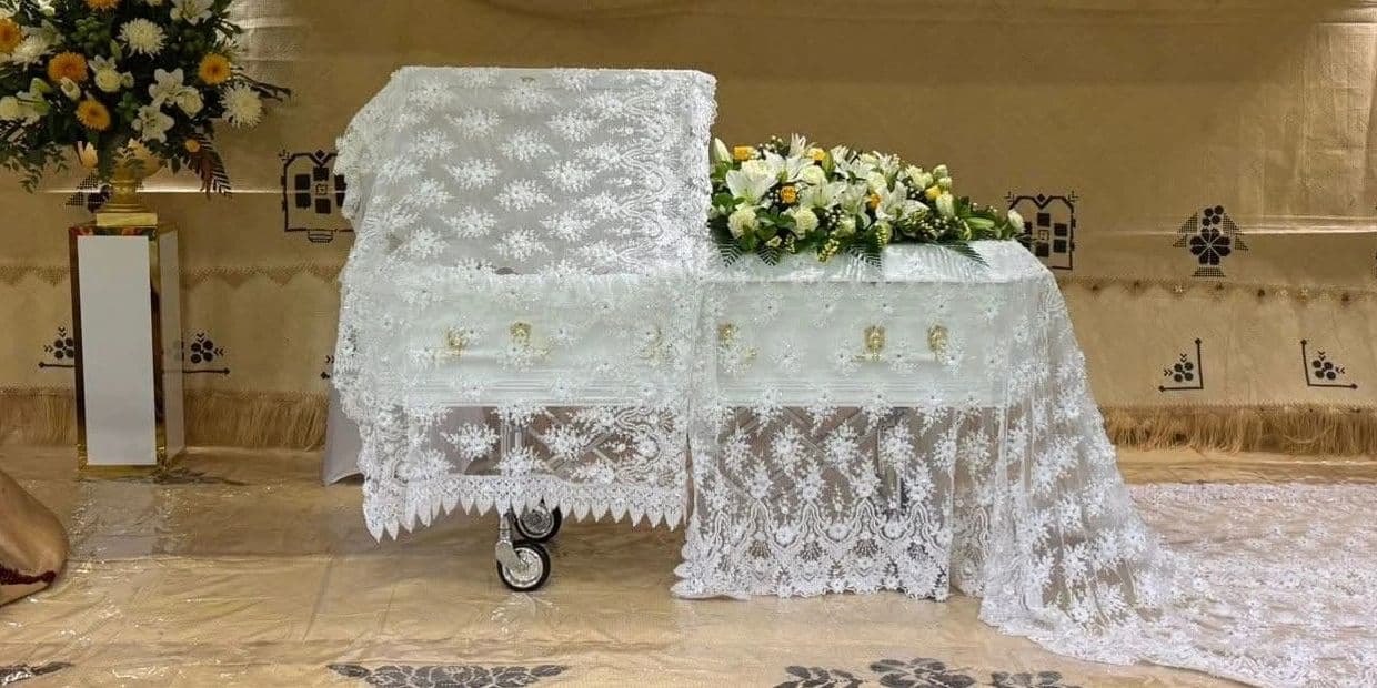 A white lace-draped casket with floral arrangements in a solemn funeral setting.
