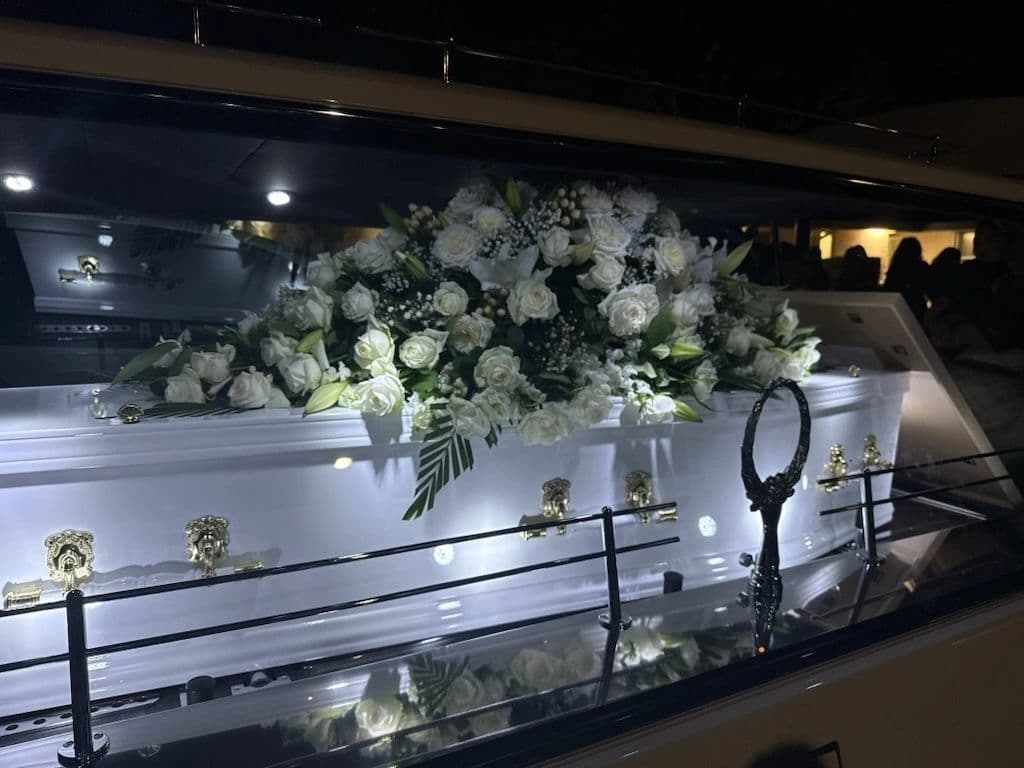 White coffin adorned with white flowers inside a hearse at night.