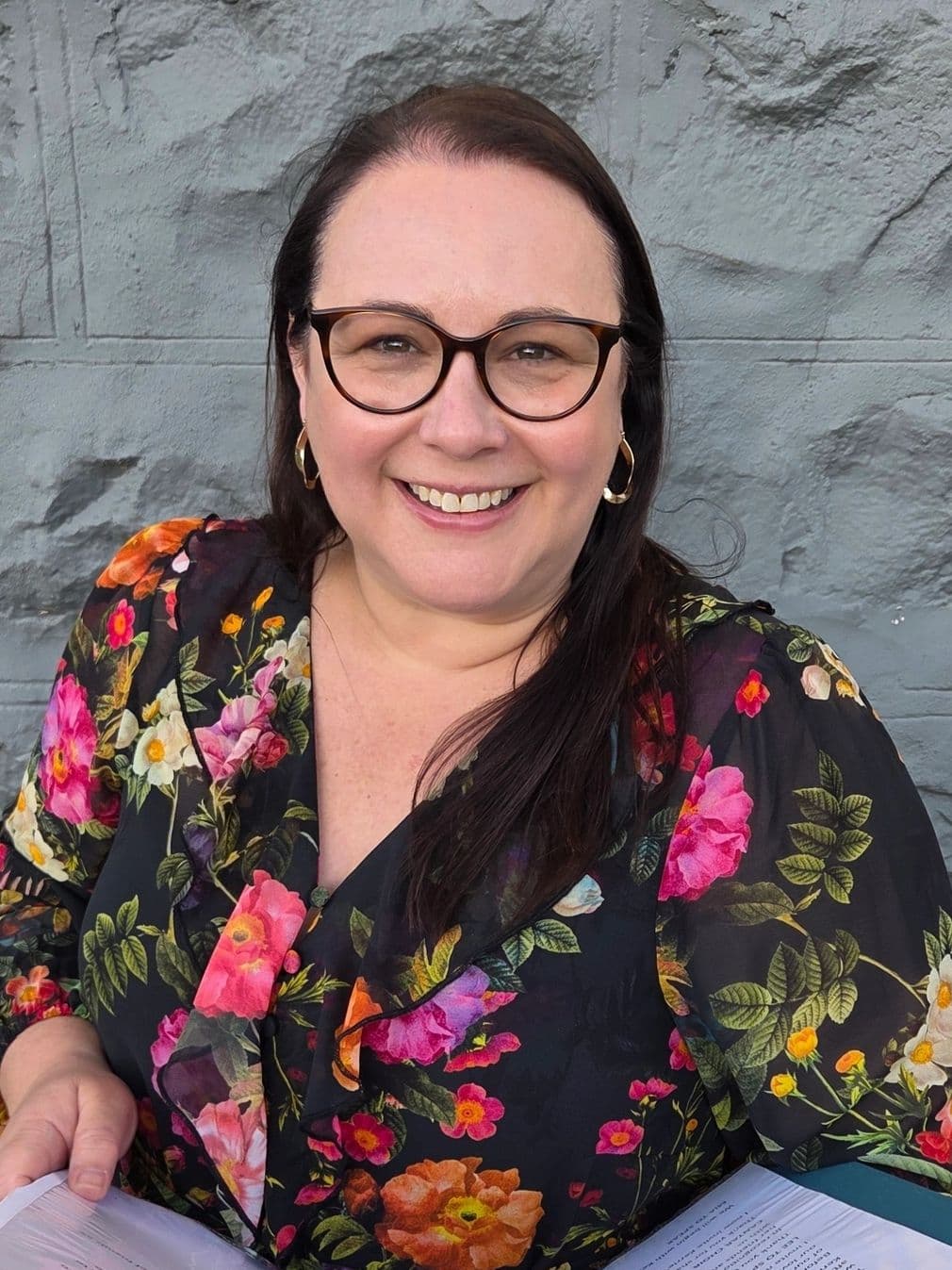 Smiling woman in glasses wearing a floral blouse, holding papers.